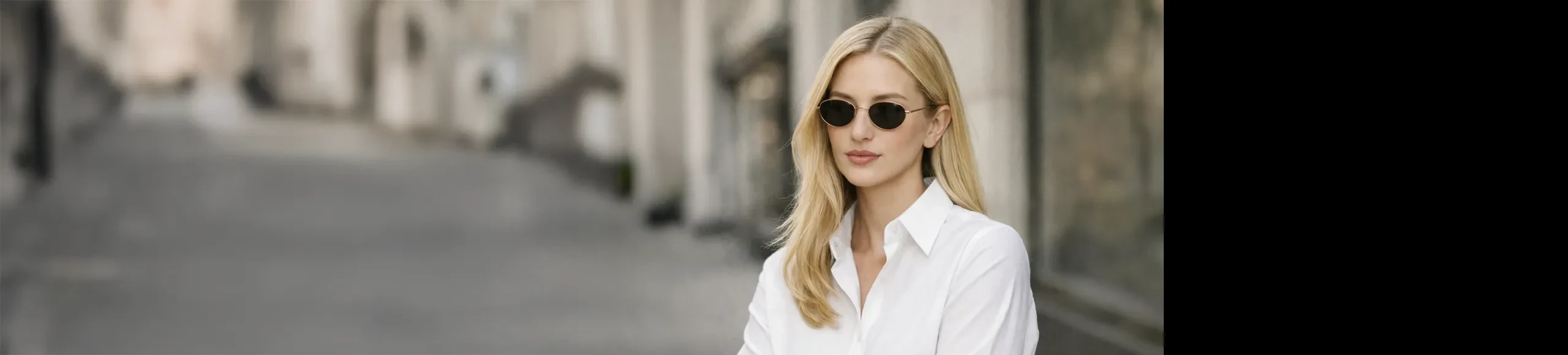 Blonde woman in a white button-down shirt and oval sunglasses standing on a blurred European street.