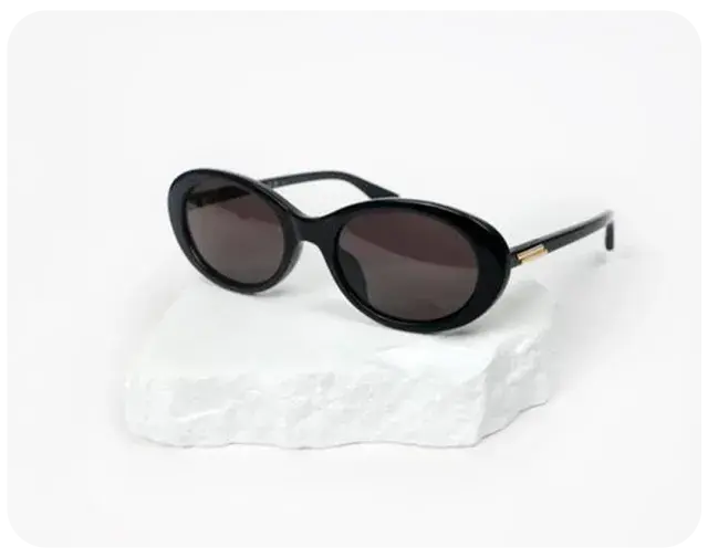 Black oval sunglasses with thick frames and gold temple accents resting on a white textured stone.