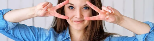 a women with brown hair in blue shirt holding up peace signs to her eyes 