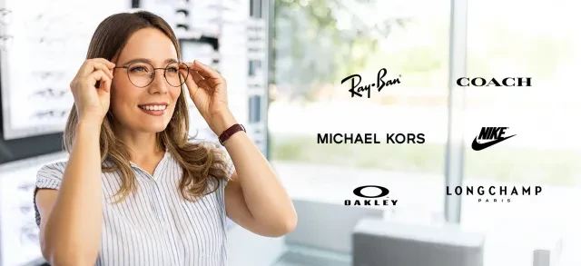 Woman trying on glasses in a bright optical store with designer brand logos displayed.