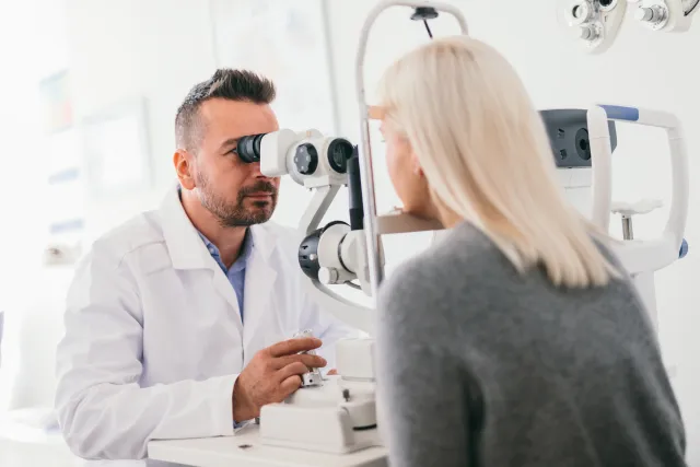 Male optometrist examines female patient's eye with an ophthalmoscope.