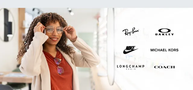Woman trying on glasses in a bright optical store with designer brand logos displayed.