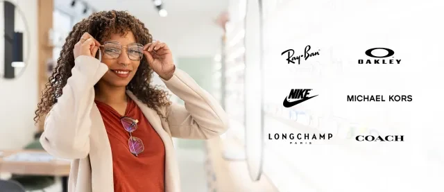 Woman trying on glasses in a bright optical store with designer brand logos displayed.